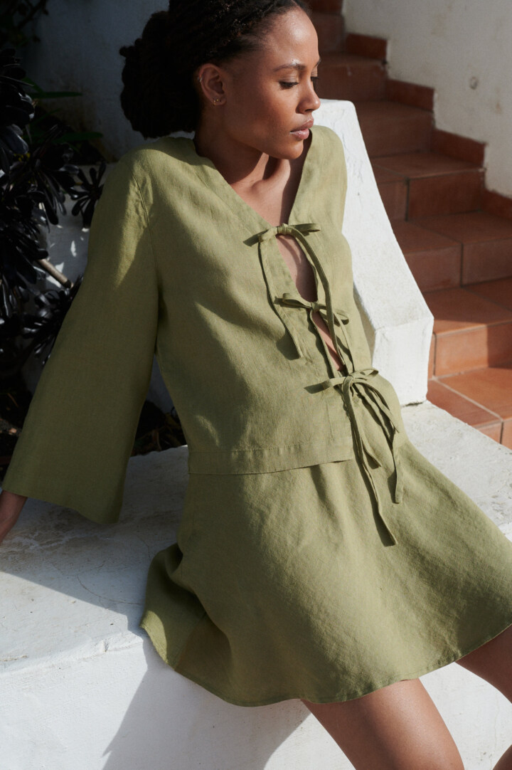 a woman is sitting in a comfortable linen set featuring a tie-up front top in olive color and a complementary short skirt in olive linen fabric