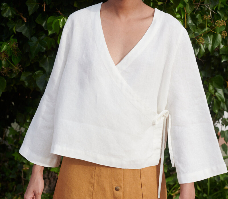 Model wearing a loose-fitting white linen top with a deep V-neck and long kimono sleeves paired with a camel linen skirt with metal snap buttons at the front