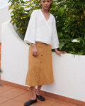 Model wearing a loose-fitting white linen V-neck wrap top and a camel color midi linen skirt with metal snap buttons