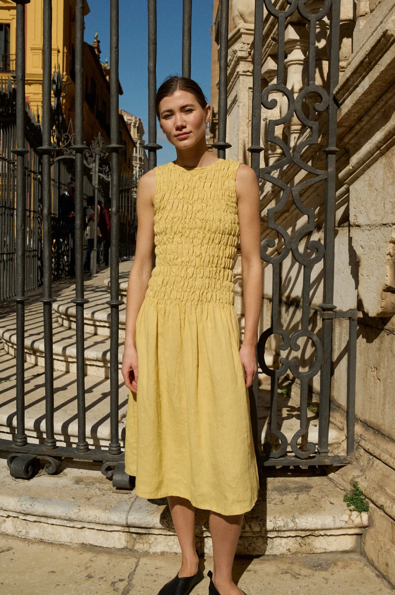 Model wearing an elegant sleeveless butter-yellow linen dress with delicate smocking detail across the bodice, transitioning into a flowing midi-length skirt for a graceful, airy silhouette
