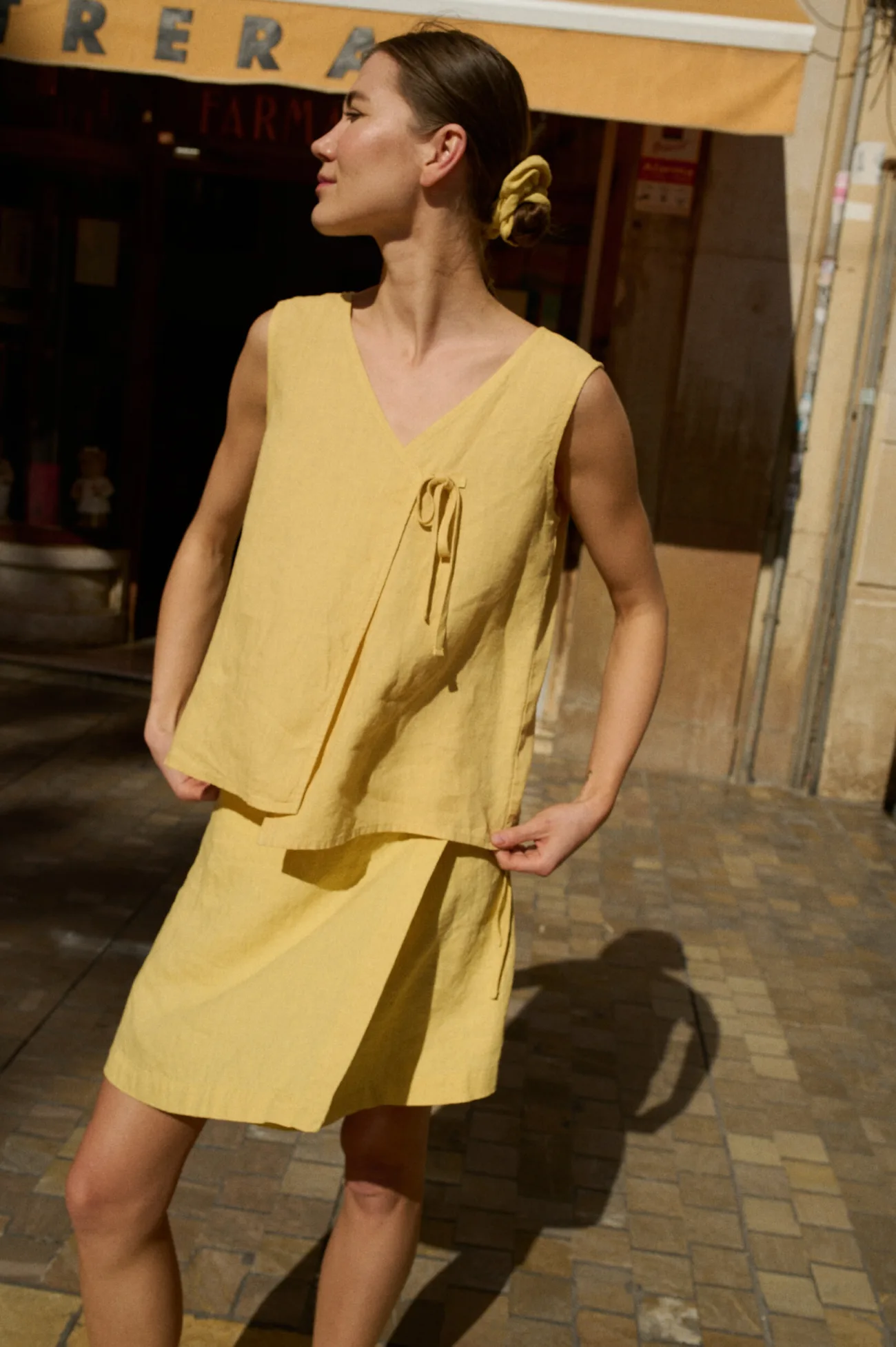 Model wearing a sleeveless, loose-fitting butter-colored linen top with an asymmetrical wrap and side tie, paired with a mini skirt in the same fabric