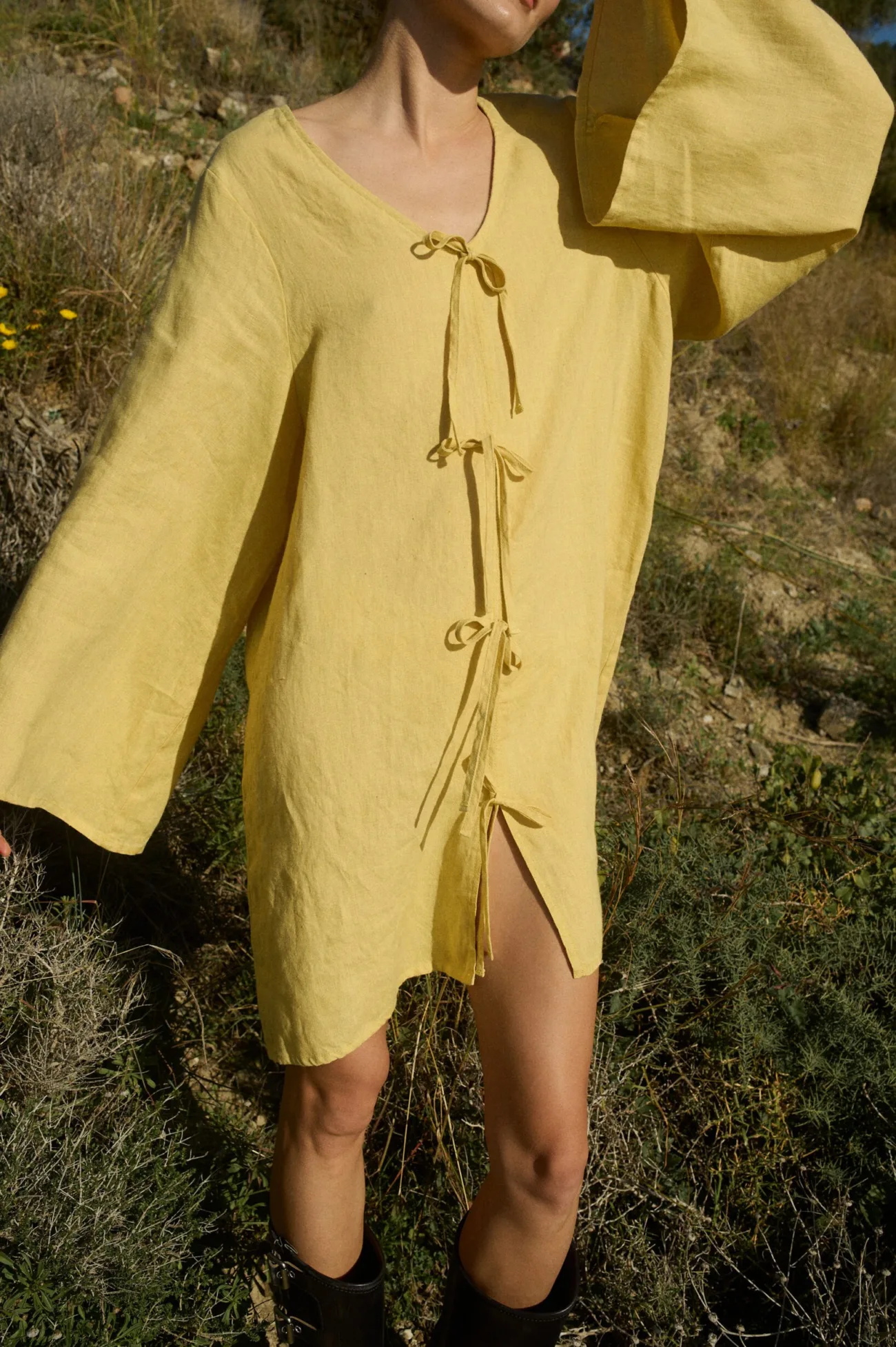 Model wearing a mini-length linen dress in butter color with wide, flowy sleeves, a deep V-neckline, and decorative tie-bows at the front, designed in a relaxed A-line silhouette and oversized fit