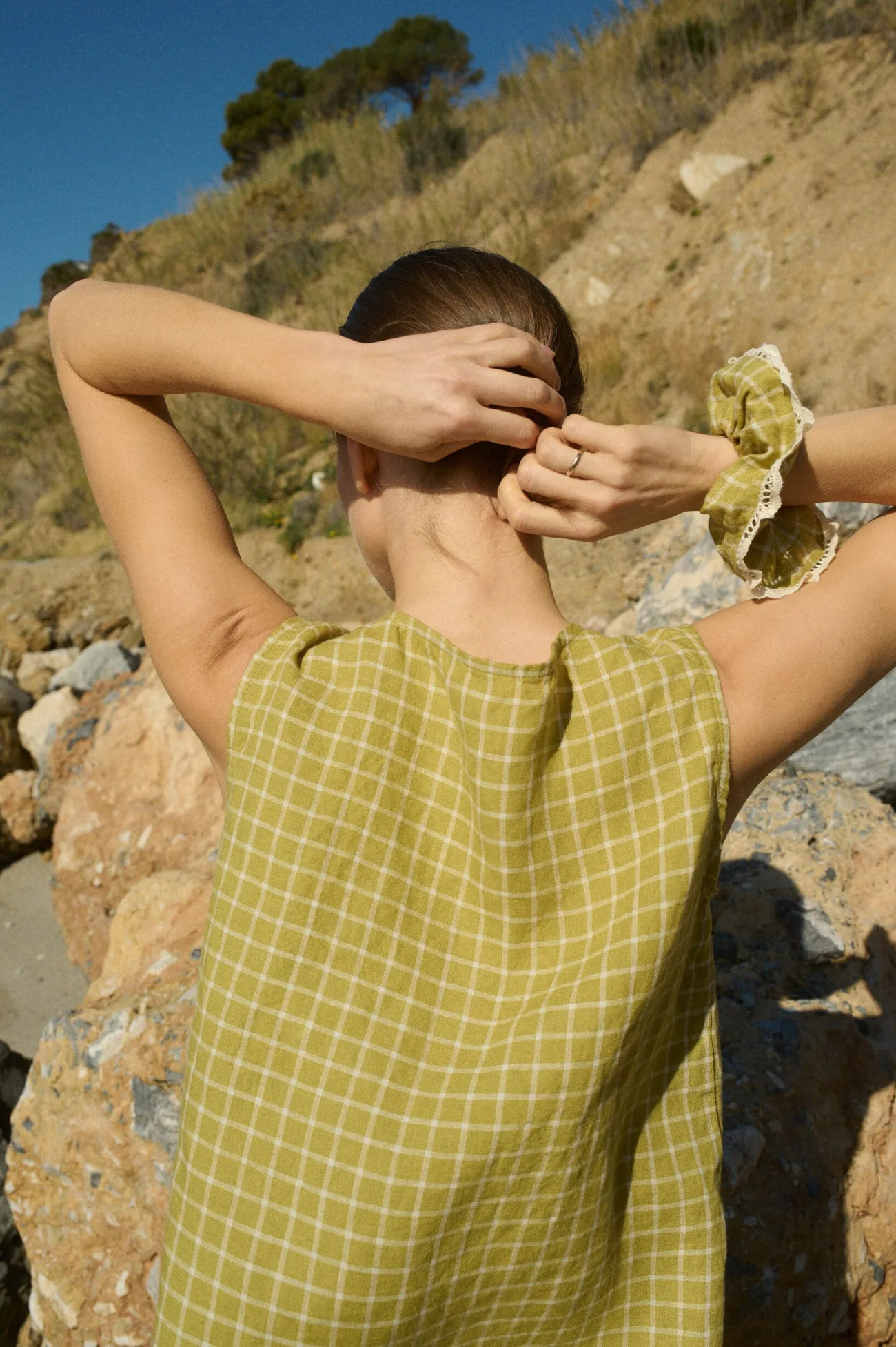 Woman wearing a pickle checks linen dress, with a scrunchie made from the same fabric featuring white lace trim, placed on her wrist as she prepares to put her hair up