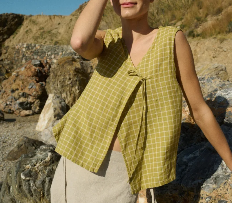 Model standing on rocky terrain, wearing a pickle checks linen sleeveless wrap top with a side tie, paired with a natural cotton blend skirt