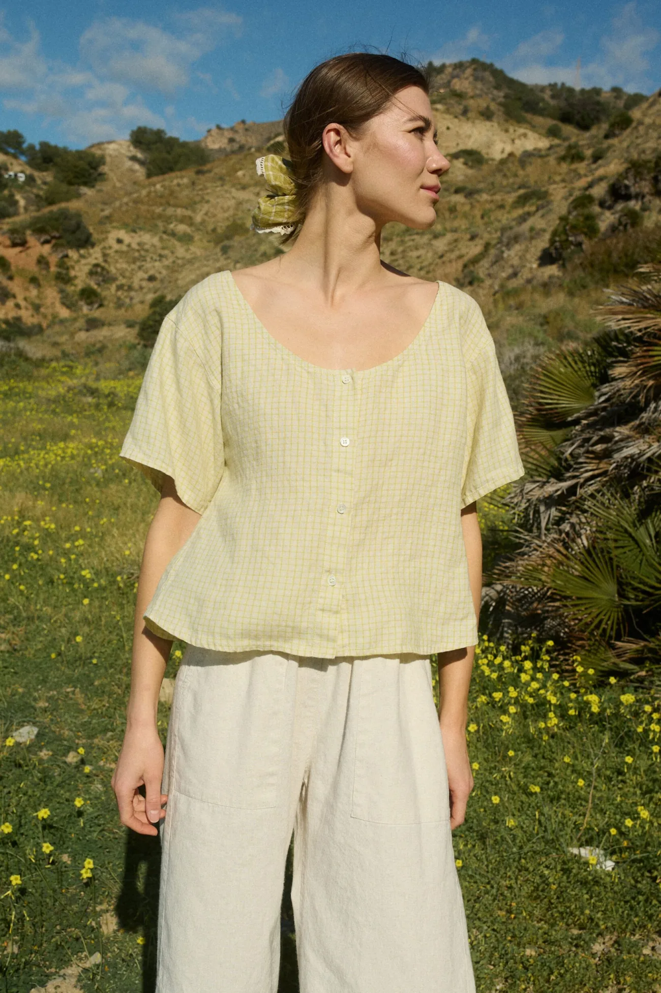 Model in a a short-sleeved, button-up top in apple checks linen, with a slightly relaxed fit, scoop neckline, and full-length buttons down the center front, paired with off-white wide-leg cotton pants featuring an elastic waistband