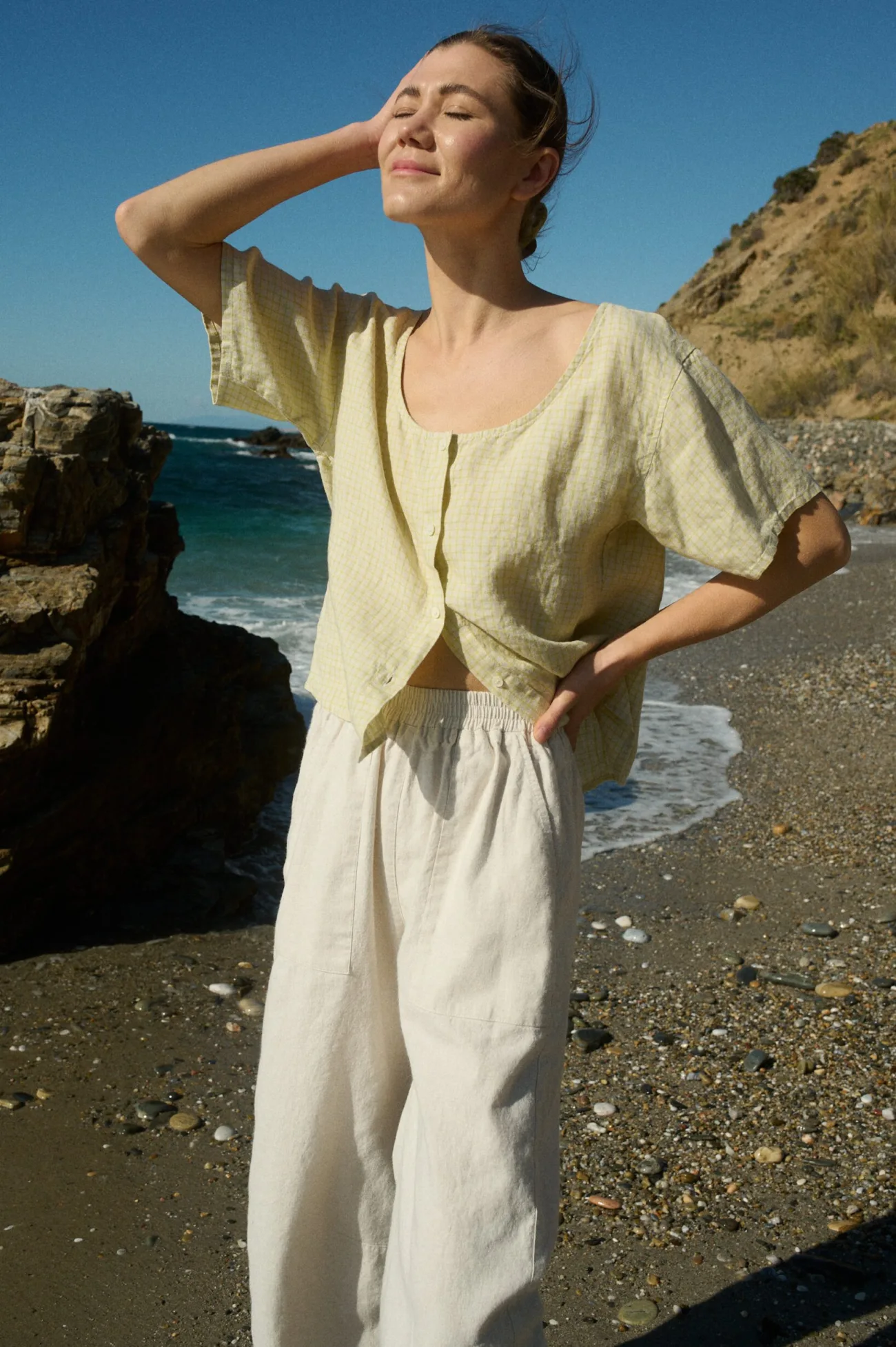 Model wearing a short-sleeved, button-up top in apple checks linen with a scoop neckline, slightly relaxed fit, and full-length buttons down the center front, paired with off-white wide-leg cotton pants with an elastic waistband