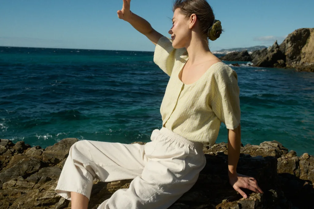 Model wearing a pale yellow checkered linen short-sleeve button-up shirt with a scooped neckline, paired with off-white wide-leg cotton blend pants with an elastic waistband and side pockets, sitting on a rock by the sea