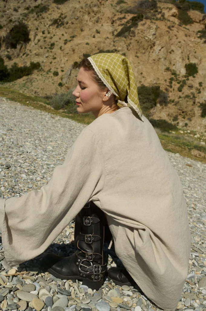 Vintage-inspired ensemble featuring an oversized natural linen tunic with flared sleeves, paired with multi-buckle black leather boots and a chartreuse checkered bandana with delicate lace trim