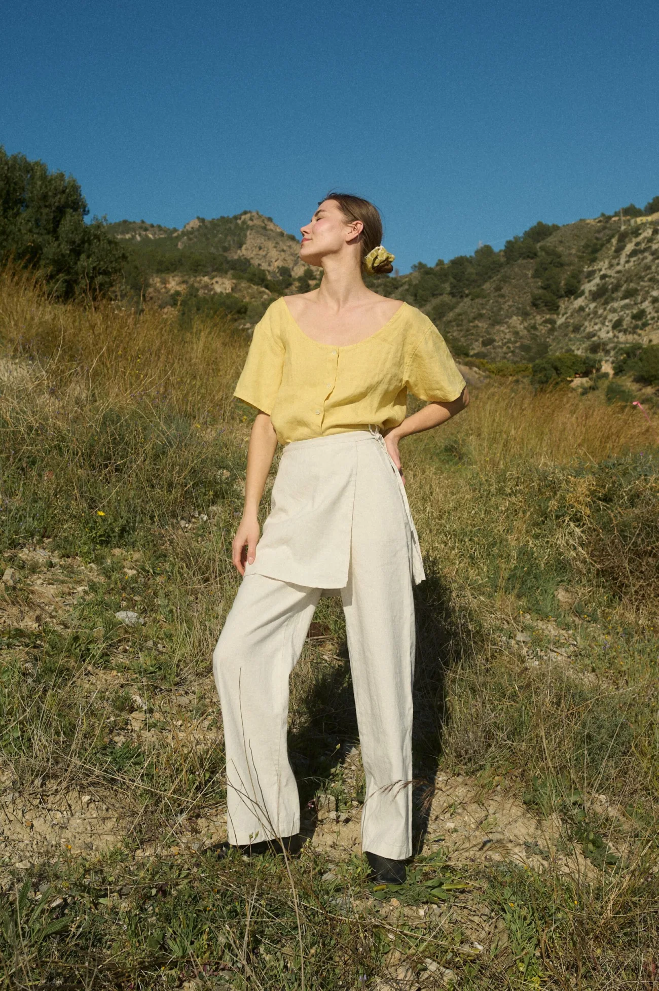 Model wearing a butter-yellow linen button-up top with a wide scoop neckline and short sleeves, tucked into white wide-leg cotton-blend pants featuring an overlapping skirt panel