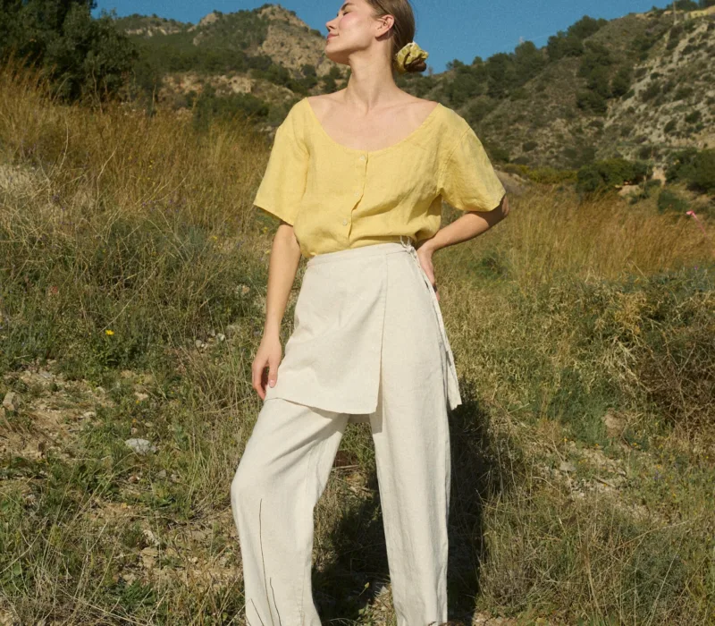 Model in nature wearing a bright yellow linen top and cream linen-cotton blend wide-leg pants with a wrap-around panel detail