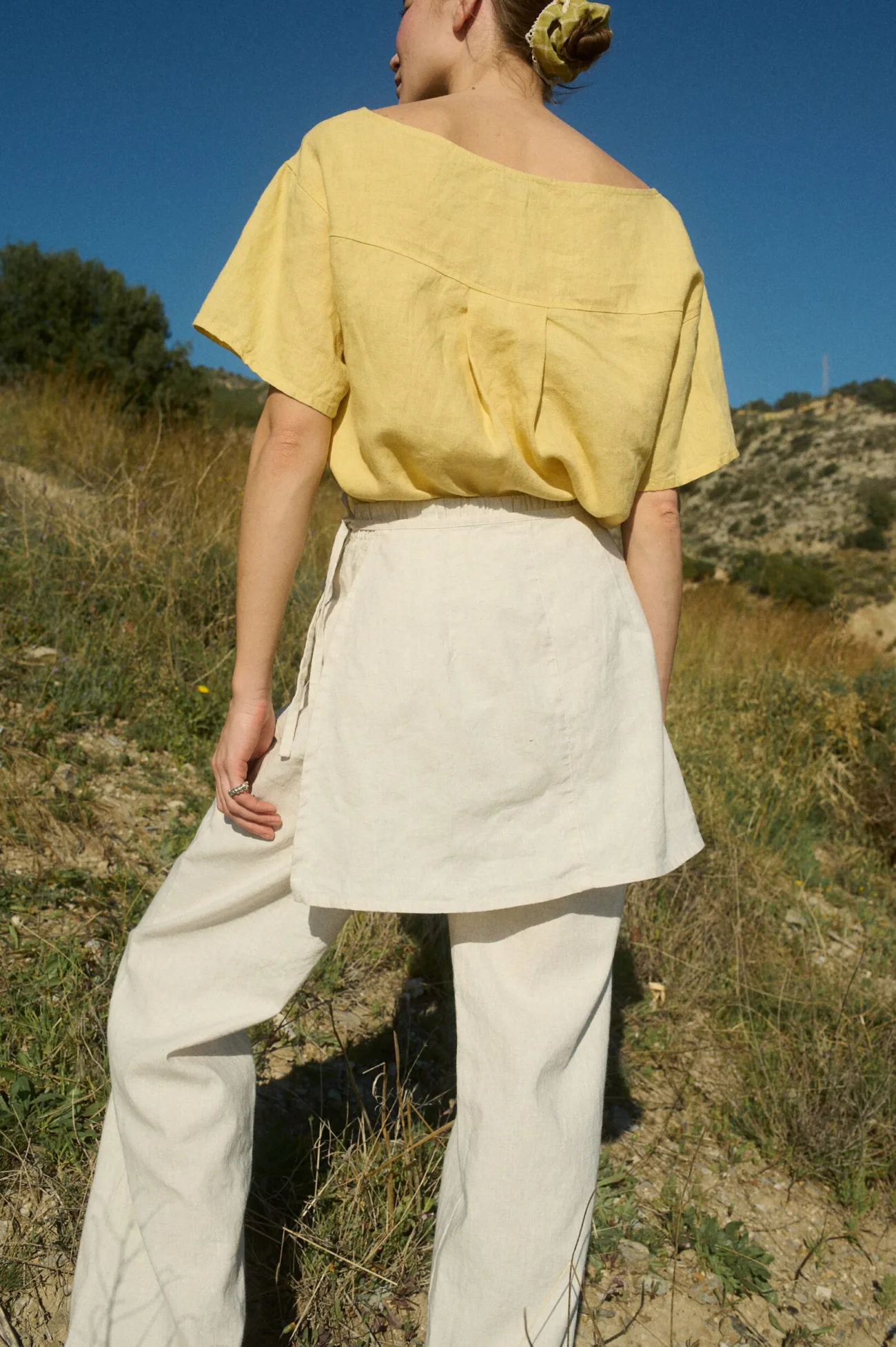 The back view showcases the yellow linen top with a wide boat neckline and elegant pleating, paired with the cream linen-cotton blend wrap and flowing wide-leg pants, set against a sunlit natural landscape