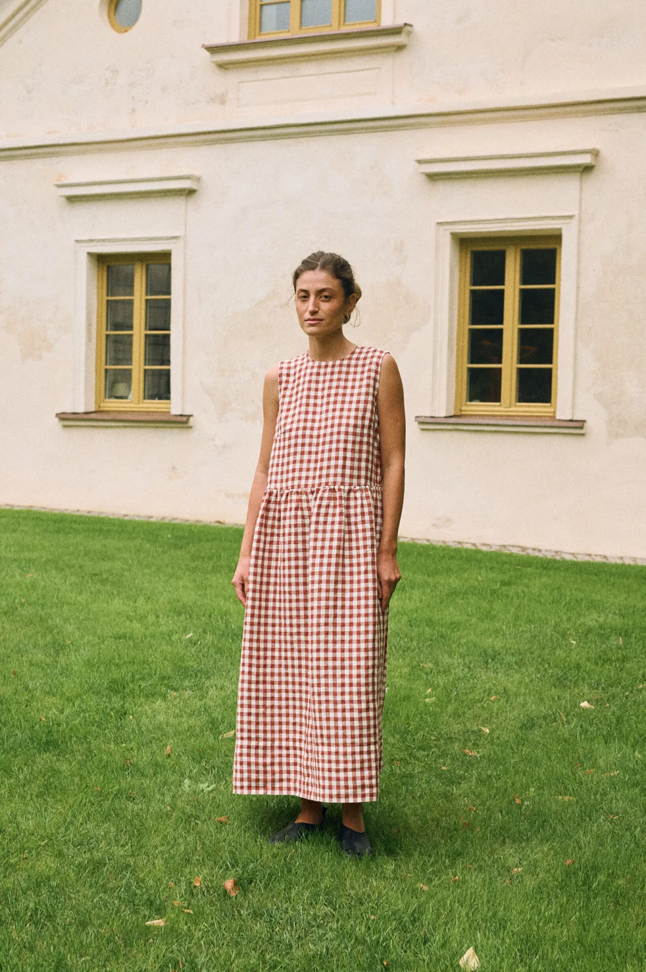 Woman dressed in a sleeveless terracotta and white gingham linen dress featuring a dropped waist and a full maxi-length skirt