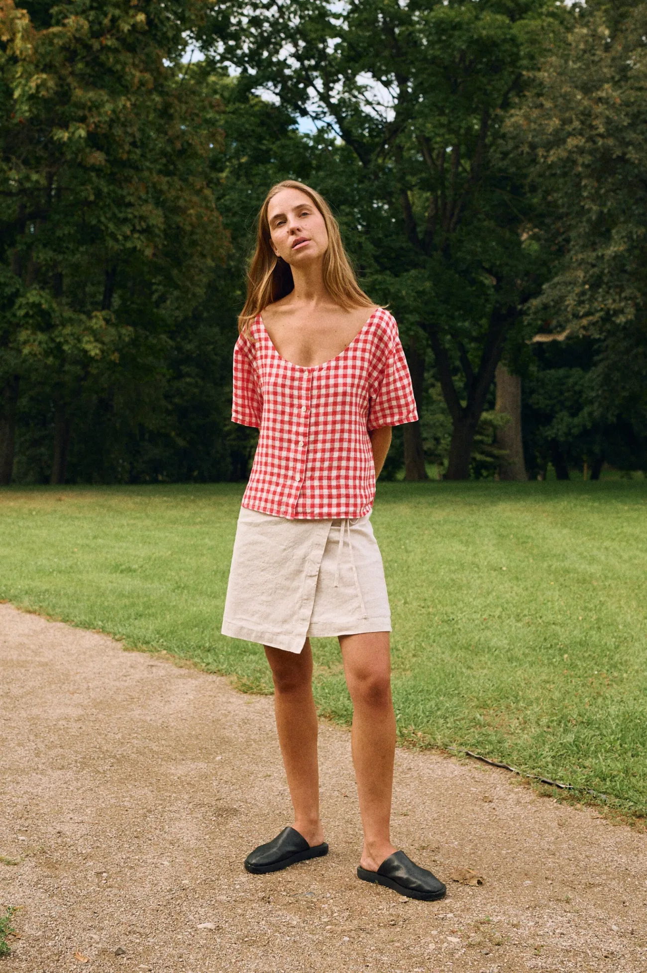 Model wearing a red gingham linen button-front top with a curved neckline and short sleeves, paired with a mini natural linen cotton blend wrap skirt