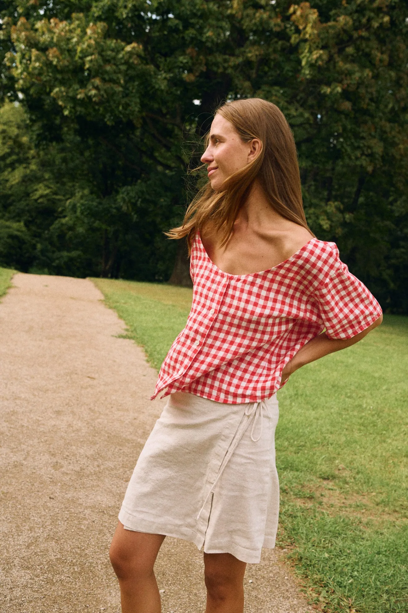 Side view of a model wearing a red gingham linen button-front top with a softly curved neckline and short sleeves, styled with a natural linen cotton blend mini wrap skirt