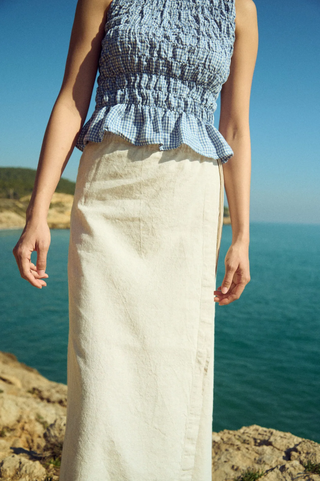 Close-up front view of a white cotton-blend wrap maxi skirt with side ties, worn with a dusty blue mini gingham linen smocked peplum top