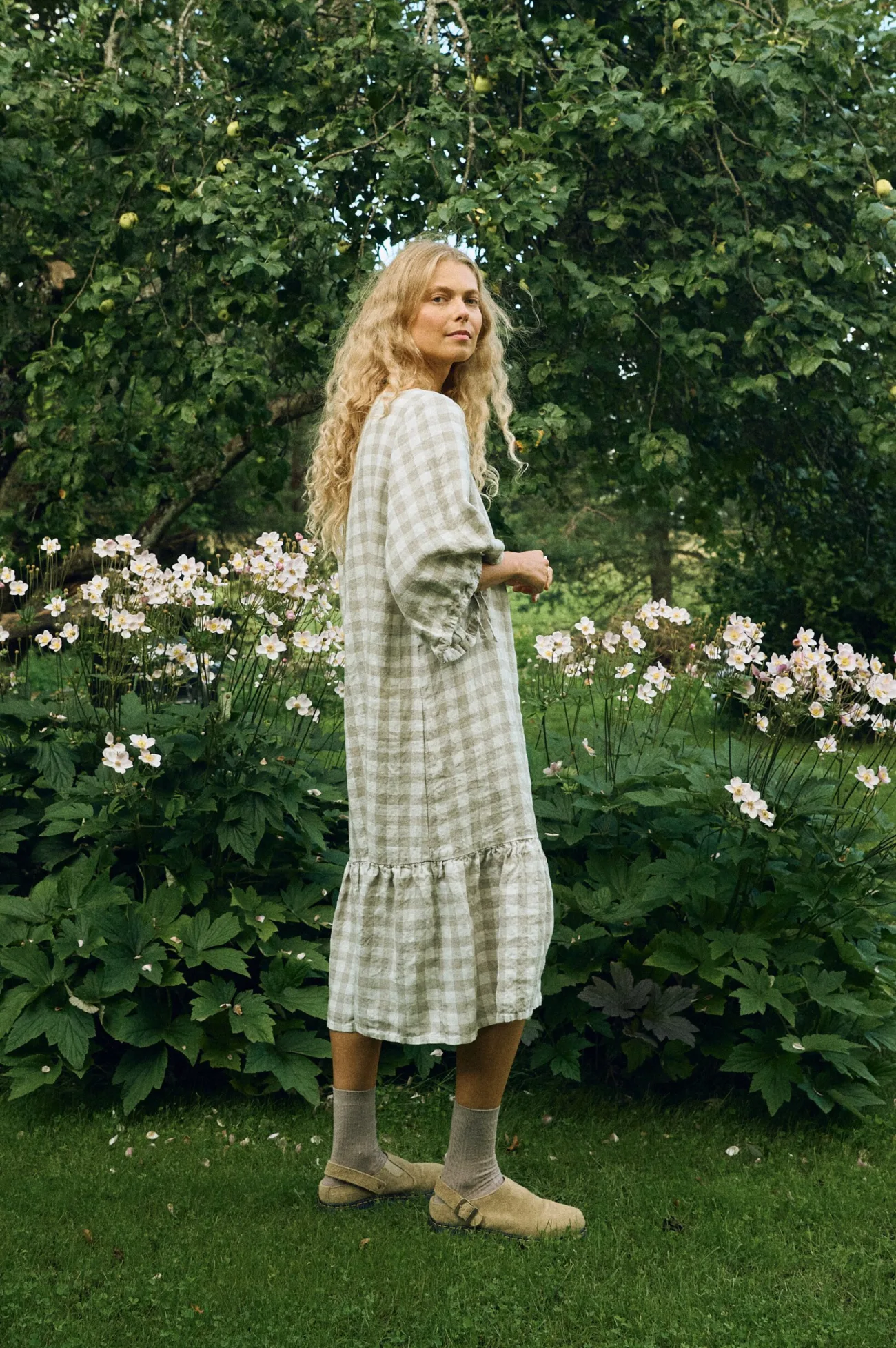 Side view of the relaxed dress in large natural gingham linen, showing its softly draped silhouette, drawstring sleeve detail, and gathered ruffle hem falling below the knee