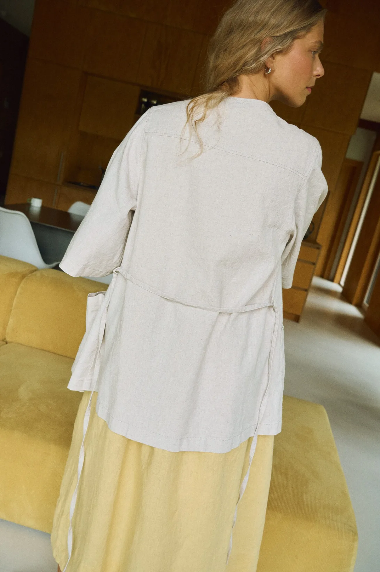 Back view of a natural linen-cotton overshirt featuring elbow-length sleeves and adjustable waist ties, worn over a soft yellow sleeveless linen midi dress