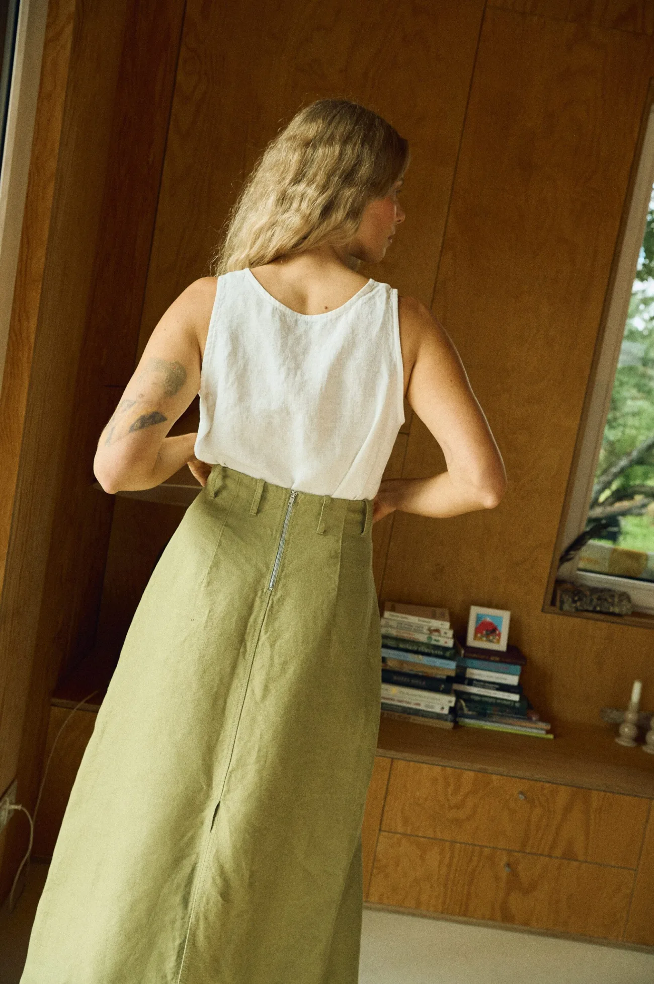 Back view of a woman wearing a white sleeveless linen top tucked into a high-waisted olive green midi skirt made from heavy linen, featuring belt loops and an exposed back zipper