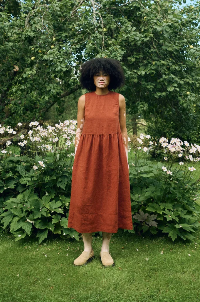 Sleeveless midi dress in terracotta linen featuring a high round neckline, fitted bodice, gathered waist, and a softly flared skirt