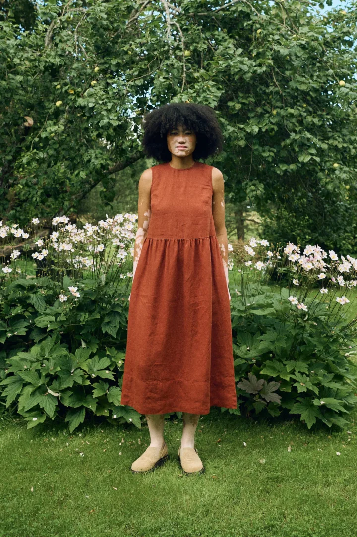 Sleeveless midi dress in terracotta linen featuring a high round neckline, fitted bodice, gathered waist, and a softly flared skirt