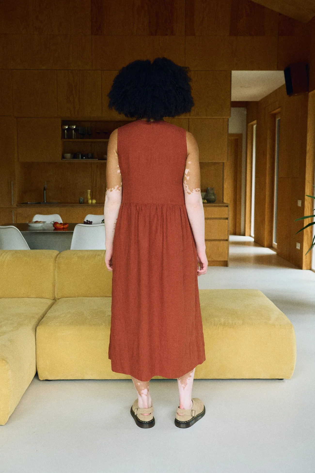 Back view of a sleeveless terracotta linen dress featuring a fitted bodice, a gathered waistline, and a softly flared midi-length skirt