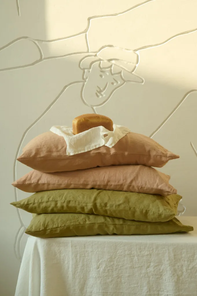 Stack of soft linen pillowcases in dusty peach and olive tones, topped with a small loaf of bread wrapped in a white linen kitchen napkin