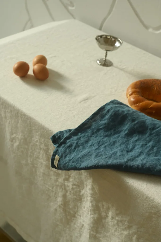 Milky white linen tablecloth with a stellar blue linen napkin draped over the edge, accompanied by three brown eggs, a silver cup, and a braided loaf of bread