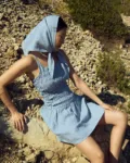 Seated side view of a dusty blue mini gingham linen mini dress with thin straps and a ruched smocked bodice, styled with a matching headscarf
