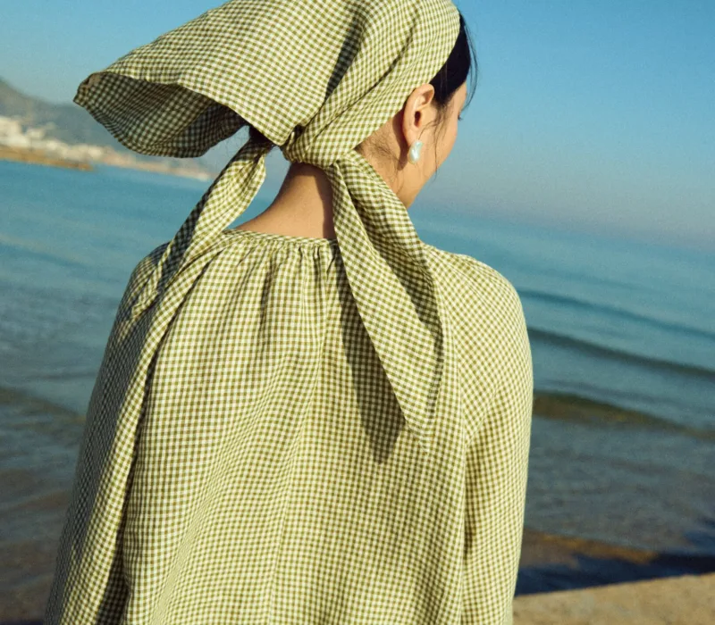 Back view of a moss green mini gingham linen triangle scarf tied at the nape, worn with a matching airy gingham blouse