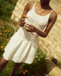 White linen mini dress with thin straps, a ruched smocked bodice, and a lightly flared skirt