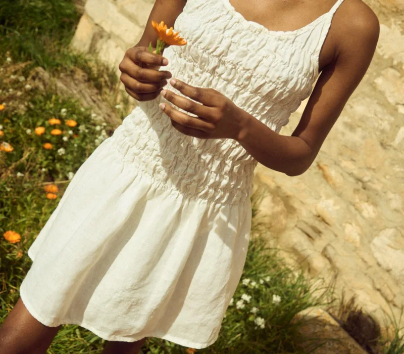 White linen mini dress with thin straps, a ruched smocked bodice, and a lightly flared skirt