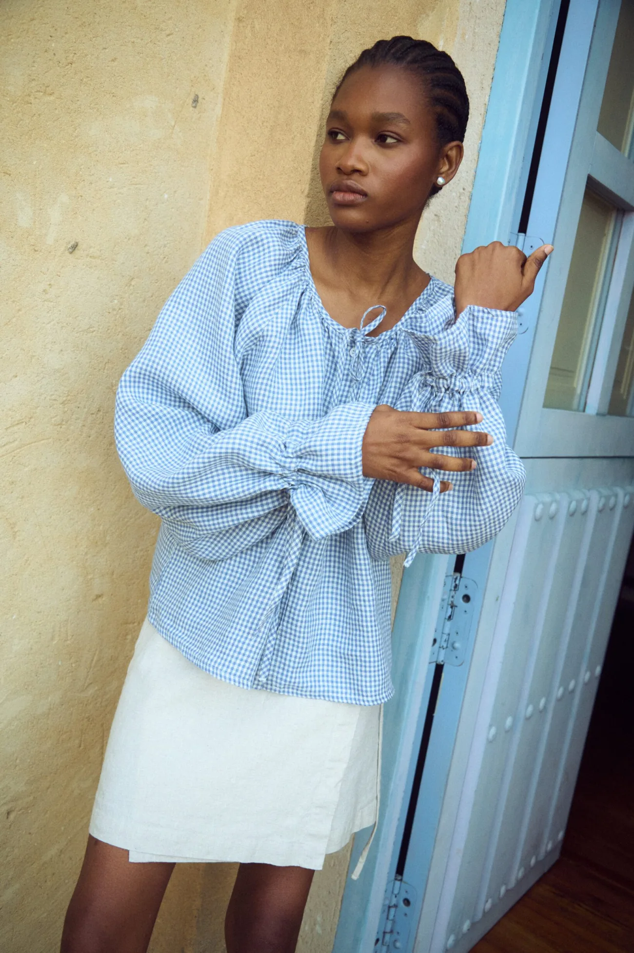 Linen top in blue gingham with relaxed fit, gathered neckline and long sleeves finished with ties, paired with a white linen mini skirt
