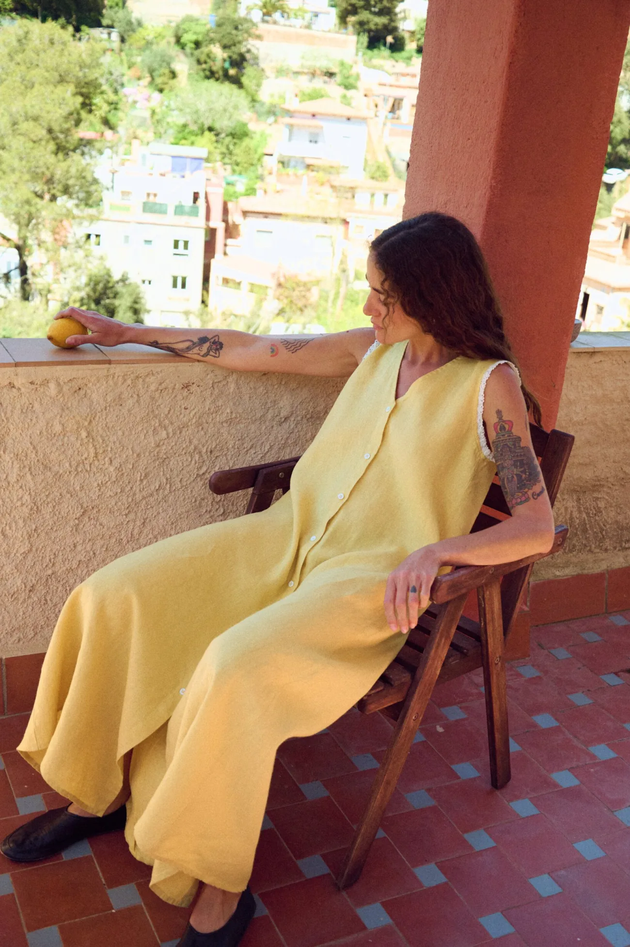 Model seated in yellow linen sleeveless maxi dress with button front, V-neck, lace-trimmed armholes, and relaxed drape