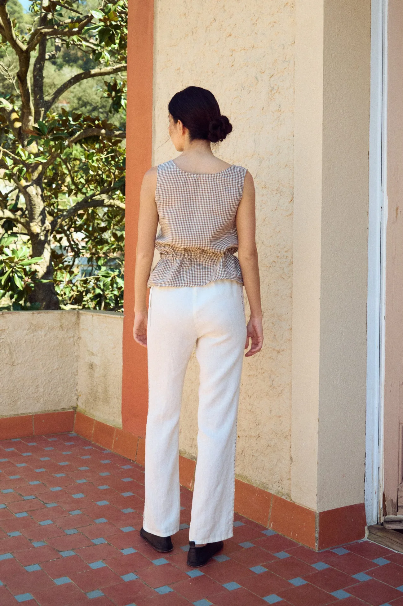 the back view of straight cut white linen pants with lace details and drawstring waist paired with peplum gingham top in brown
