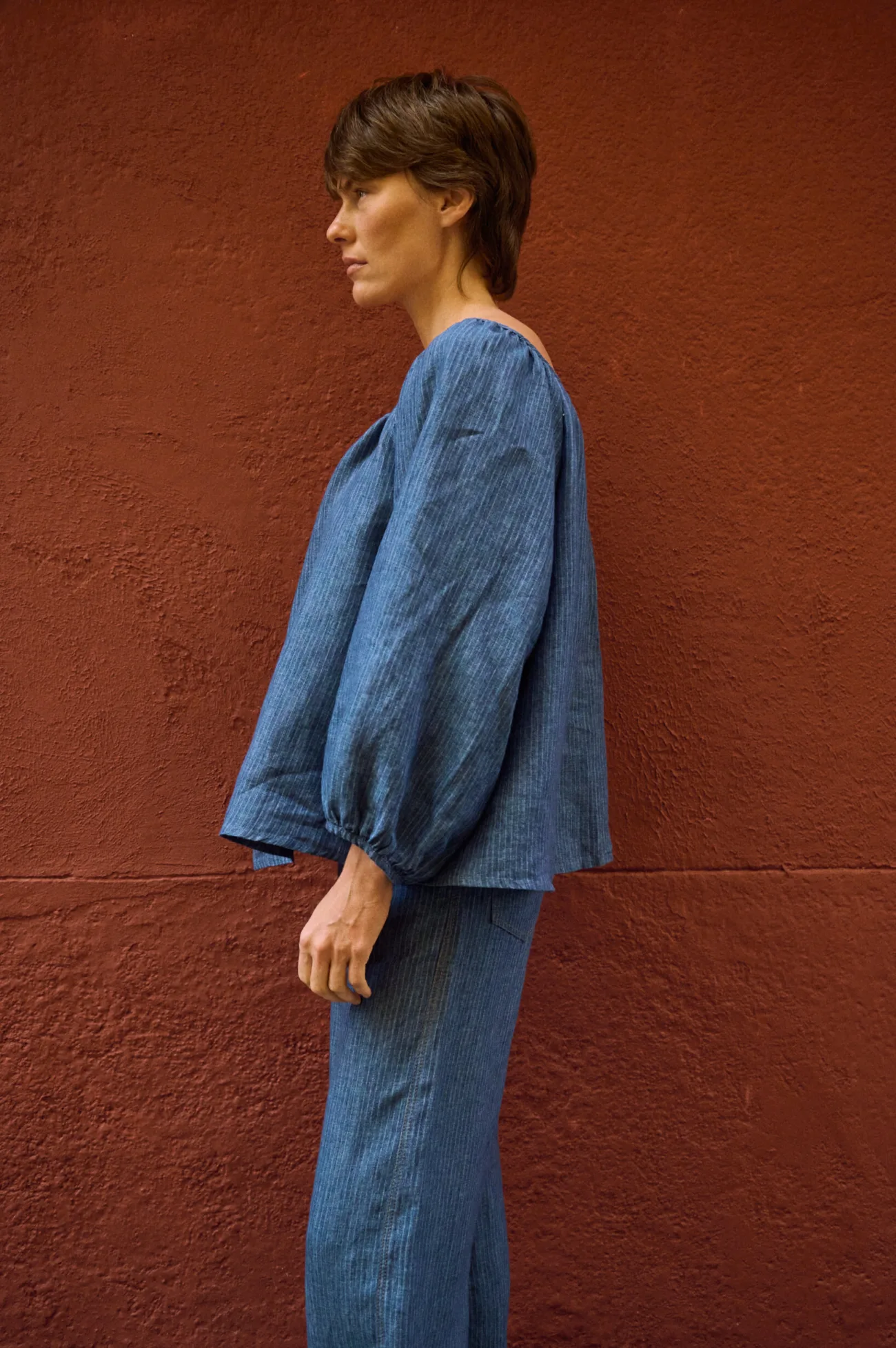 Side view of a blue striped linen blouse with a relaxed shape and voluminous sleeves, paired with matching linen pants