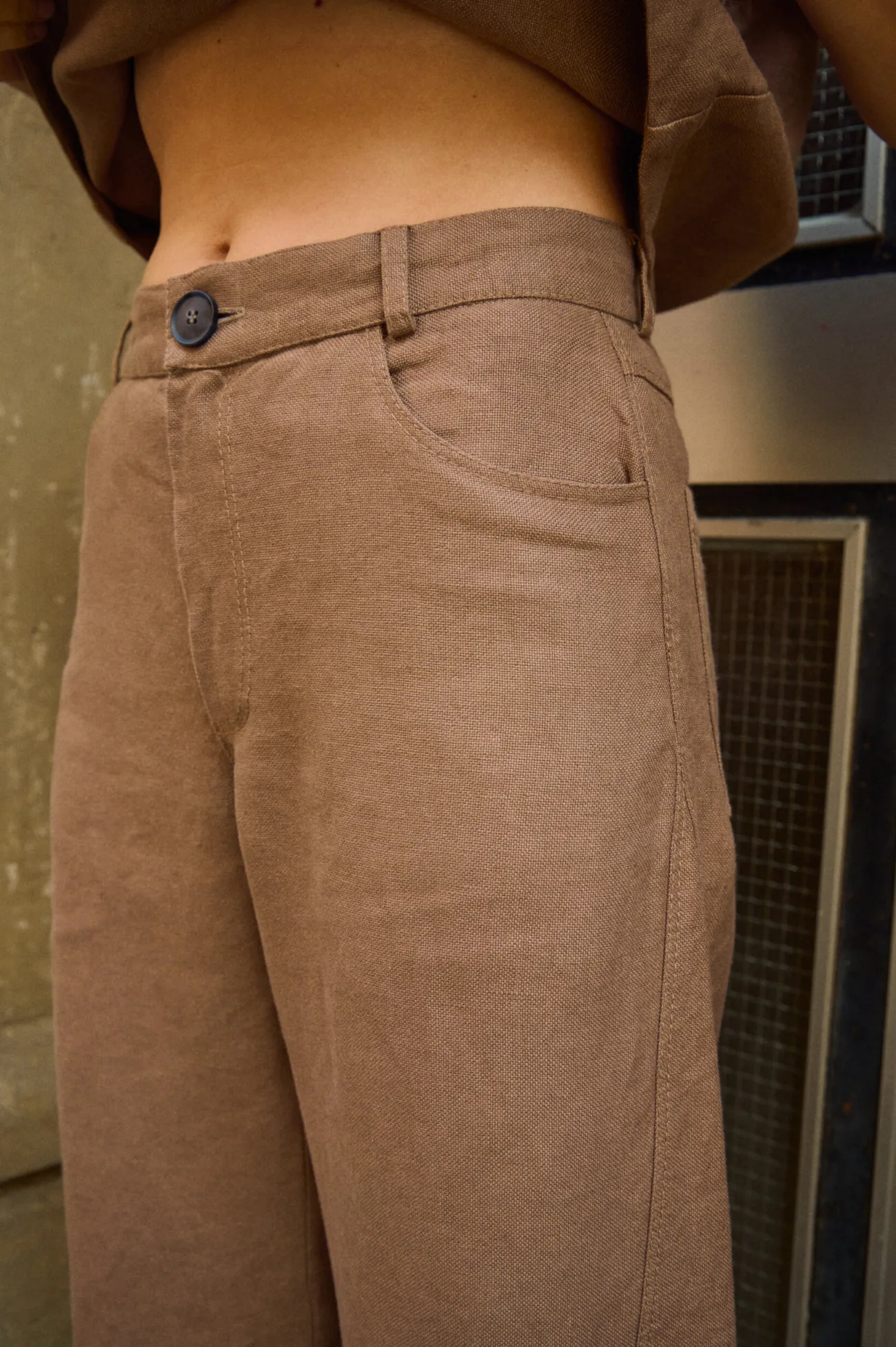 Close-up of brown heavy linen trousers showing the waistband, dark button, belt loops and curved front pocket